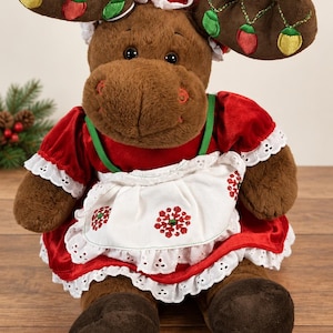May include: A brown plush moose toy wearing a red velvet dress with a white eyelet lace apron featuring red snowflake embroidery. The moose has antlers decorated with embroidered colorful light bulbs and wears a matching red velvet bonnet.