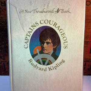 May include: A vintage hardcover book titled "Captains Courageous" by Rudyard Kipling. The cover features a portrait of a young person and a ship's wheel with a rainbow. The book is from "A New Thrushwood Book".