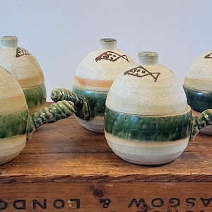 May include: A collection of five ceramic lidded pots with handles. The pots are cream-colored with a green band around the middle and a fish design on the lid. The handles are a twisted green design. The pots are displayed on a wooden surface.