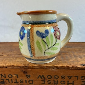 May include: A ceramic pitcher with a cream-colored base, adorned with blue and maroon floral designs and green leaves. The pitcher has a brown rim and handle. The background is a wooden surface.