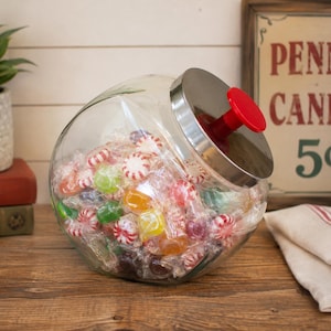 May include: A clear glass jar with a slanted opening and a silver metal lid featuring a bright red knob handle. The jar is filled with assorted individually wrapped candies in various colors including red, green, yellow, and white striped patterns.