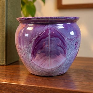 May include: A rounded ceramic planter featuring a glossy finish with a marbled pattern in shades of deep purple, violet, and light lavender. The vessel has a wide, slightly flared rim and a smooth, curved body with a distinct central design.