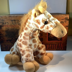 May include: A plush giraffe toy with a white and brown spotted pattern. The giraffe has a tan snout, ears, and feet. The toy is sitting in front of a wooden crate.