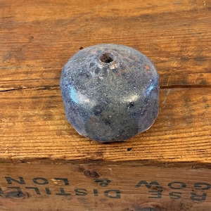 May include: A weathered, blue-gray, rounded object with a hole at the top, resting on a wooden surface. The object has a textured appearance and a slightly glossy finish. The wood has a warm, brown tone.