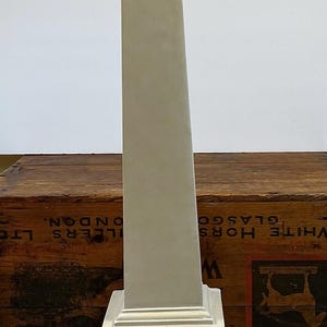 May include: A tall, light gray obelisk sculpture featuring a pointed pyramidal top, a slender tapering shaft, and a stepped square base. The object has a smooth, matte finish and stands vertically as a singular, solid decorative architectural piece.