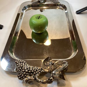 May include: A rectangular silver serving tray with decorative grape and leaf handles. A green apple sits in the center of the tray, reflecting on the shiny surface. The tray has a polished finish, suitable for serving food or displaying items.