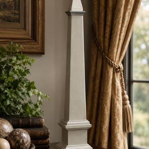 May include: A tall, light gray obelisk sculpture featuring a square base, a tapered four-sided shaft, and a pointed pyramidal top. The object has a smooth, matte finish and stands vertically as a singular decorative piece on a flat surface.