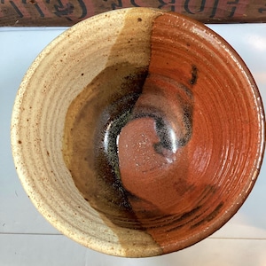 May include: A handmade ceramic bowl with a two-tone design. The left side is a light beige with brown speckles, while the right side is a reddish-brown. The bowl has a glossy finish and a spiral pattern.