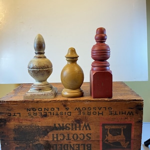 May include: Three decorative wooden finials in varying shapes and colors. The finials are placed on a wooden crate with text that reads "White Horse Distillers" and "Scotch Whisky". The finials are painted in shades of brown and red.