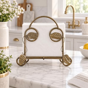 May include: A bronze-toned metal napkin holder featuring a curved handle, circular side accents, and decorative leaf motifs at the base and sides. The structure holds a stack of square white paper napkins in an upright position on a flat surface.