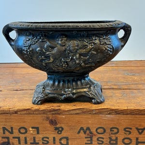 May include: A dark gray cast iron planter with a pedestal base. The planter features relief carvings of cherubs, grapes, and vines. It has two handles on either side. The planter is sitting on a wooden crate.