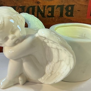 May include: A white ceramic angel figurine with a small planter. The angel is in a seated position, with its head resting on its knees. The planter is a light green color and is attached to the angel's wing.