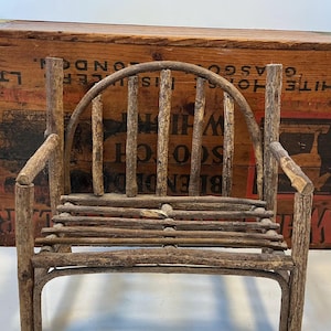 May include: A miniature rustic bench crafted from natural twigs and branches. The bench features a curved backrest with vertical supports, armrests, and a slatted seat. The wood has a natural, weathered appearance, showcasing the texture of the bark.