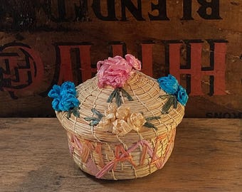 May include: A small round woven basket with a lid, featuring natural tan straw fibers. The lid is decorated with three clusters of artificial flowers in pink, blue, and yellow. The base of the basket is accented with woven pink and orange ribbon.