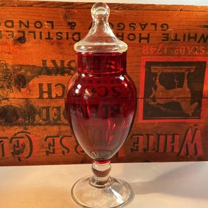 May include: A ruby red glass apothecary jar with a clear glass lid and base. The jar has a bulbous body and a slender stem, and it sits on a round base. The background is a wooden crate with faded lettering.