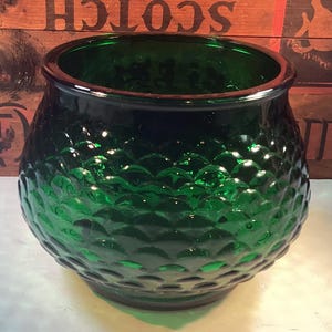 May include: A dark green glass bowl with a textured, dimpled surface. The bowl has a rounded shape with a wide opening and a thick rim. The glass is a deep emerald color, reflecting light.
