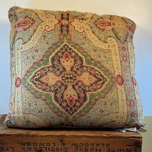 May include: Square decorative pillow with an intricate, ornate pattern in shades of green, red, and beige. The pillow has a central medallion design and a textured appearance, suggesting a woven fabric. The edges are trimmed with a rope-like detail.