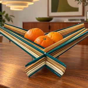 May include: A collapsible fruit bowl constructed from intersecting wooden slats in natural light wood and teal finishes. The geometric, X-shaped structure holds three bright orange citrus fruits nestled within its open, angular frame.