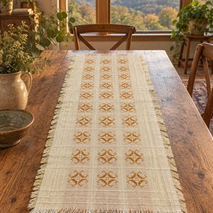 May include: A rectangular cream-colored table runner featuring a repeating geometric pattern in mustard yellow. The runner is crafted from a woven fabric with a grid-like texture and finished with frayed, fringed edges along all four sides.