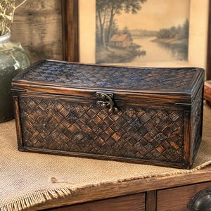 May include: A rectangular dark brown wooden box with a woven texture pattern on the lid and front panels. The box features a hinged lid secured by a small, centered metal latch. The finish is a deep, stained wood tone with visible grain.