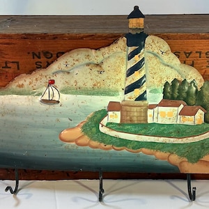 May include: A decorative wooden wall hanging featuring a lighthouse scene. The lighthouse has a black and white striped tower, with a light at the top. The scene includes a sailboat, houses, and trees. Two black metal hooks are attached to the bottom.