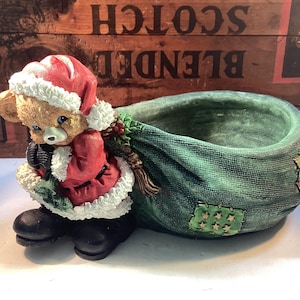May include: A Christmas-themed figurine featuring a teddy bear dressed as Santa Claus, carrying a green sack. The bear wears a red coat and hat with white trim. The sack has decorative patches.