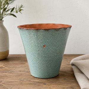 May include: A tapered ceramic planter featuring a light turquoise exterior with a dark, intricate crackle glaze pattern. The interior rim and base reveal a contrasting terracotta orange clay color. A small, circular gold-toned accent is centered.