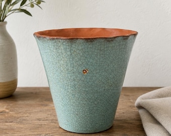 Puede incluir: Una maceta de cerámica cónica con un exterior de color turquesa claro y un patrón de esmalte craquelado oscuro e intrincado. El borde interior y la base revelan un color arcilla naranja terracota contrastante. Un pequeño acento circular dorado está centrado.