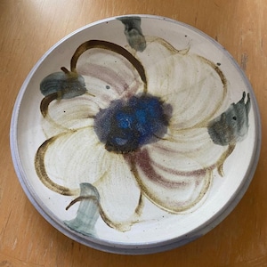 May include: A round ceramic plate with a cream-colored base featuring a hand-painted floral design. The center of the flower is a deep blue, surrounded by petals outlined in brown and green brushstrokes. The plate has a slightly raised rim.