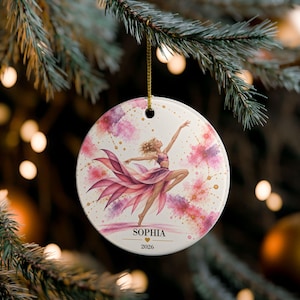 May include: A round white ceramic ornament featuring a watercolor illustration of a dancer in a flowing pink dress. The design includes pink and purple splashes, gold dots, and the text SOPHIA above a small heart and the year 2026 in black font.
