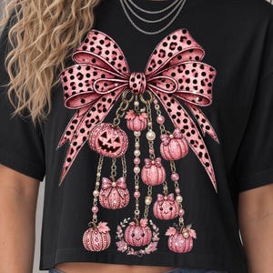 Peut inclure: Un t-shirt noir orné d'un grand nœud rose à motif léopard. Suspendues au nœud, plusieurs citrouilles roses, dont certaines avec des visages, sont reliées par des chaînes dorées et des perles. Le motif présente des nuances de rose et d'or.