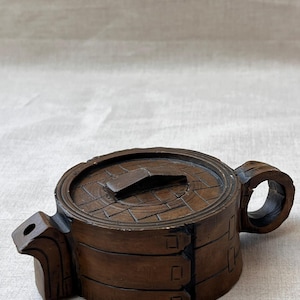 May include: A dark brown wooden teapot featuring a cylindrical body with carved horizontal bands and vertical slats. The lid has a woven-style texture with a central rectangular handle, and the side includes a curved spout and a circular handle.