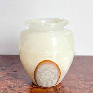 May include: A small, rounded vase crafted from translucent, pale cream-coloured stone. The vessel features a smooth, polished surface with a distinct, oval-shaped inclusion near the base that displays a textured white centre and a brown rim.