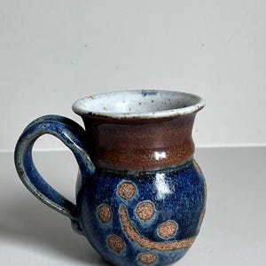May include: A ceramic mug with a rounded body and a curved handle. The upper portion is a smooth brown glaze, while the lower section features a deep blue mottled glaze with exposed tan clay circular and curved patterns throughout the surface.