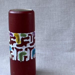 May include: A cylindrical ceramic vase with a matte dark red finish. The centre of the vase features a wide, horizontal band of raised white glaze with abstract, flowing shapes in yellow, green, purple, and blue accents painted within the white.