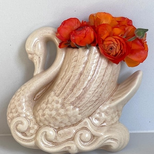 May include: A cream-coloured ceramic swan-shaped vase with a glossy finish and textured feather details. The base features stylised wave patterns. A cluster of vibrant orange ranunculus flowers is arranged inside the swan's back opening.
