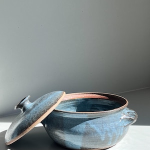 May include: A round ceramic pot with a lid, featuring a glossy blue glaze over a dark clay body. The pot has two small side handles and a matching lid with a central knob. The interior and rim reveal the natural reddish-brown clay texture.