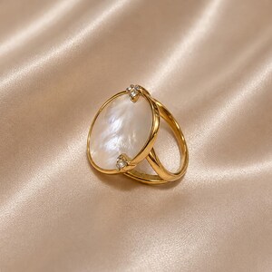 May include: A gold-toned ring featuring a large, oval-shaped white mother-of-pearl inlay. The metal band splits into two sections as it reaches the setting, which is accented by two small, clear, round-cut stones at the top and bottom edges.