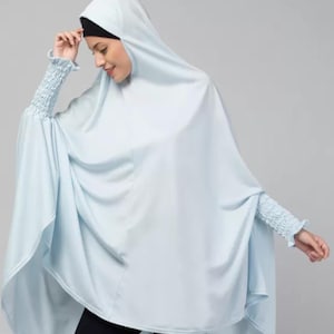 May include: A light blue hooded poncho featuring long sleeves with gathered, smocked cuffs. The garment has a loose, flowing silhouette that drapes over the torso, providing full coverage. It is worn over a black head covering and black pants.