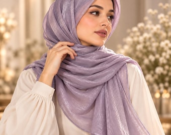 May include: A shimmering lavender headscarf draped elegantly around the head and shoulders, featuring a lightweight, textured fabric with a subtle metallic sheen. The scarf is worn over a plain, long-sleeved cream-colored blouse with buttoned cuffs.