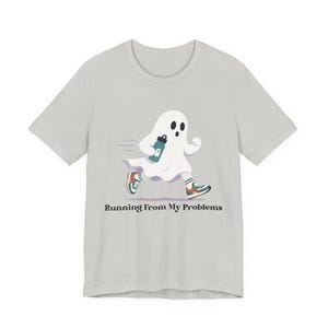 May include: A light grey t-shirt featuring a graphic of a white ghost running while holding a teal water bottle. The ghost wears colourful trainers and striped socks. Below the graphic, the text reads, Running From My Problems in black.