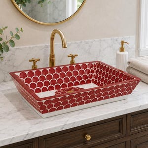 May include: A rectangular vessel sink featuring a repeating red and white scalloped pattern on the interior and exterior surfaces. The sink is equipped with a gold-toned faucet and two matching cross-handle knobs, set against a white marble countertop.