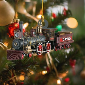 May include: A hanging Christmas tree ornament shaped like a vintage steam locomotive. The train is black with red wheels, gold accents, and a maroon tender featuring the name DAVID in white capital letters. It hangs from a gold string.