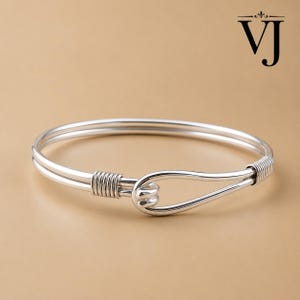 May include: A silver-toned bangle bracelet featuring a double-wire design that converges into a hook-and-eye style clasp. The ends are secured with coiled wire detailing. The letters VJ are printed in black in the upper right corner of the frame.