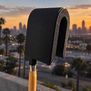 May include: A black, U-shaped, curved tool head attached to a light-coloured wooden handle. The tool features a smooth, matte black finish on its exterior surface and a metallic silver connector where the head meets the wooden shaft.