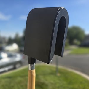 Op de afbeelding: Een zwarte U-vormige gereedschapskop bevestigd aan een lichtgekleurde houten steel. Het gereedschap heeft een gladde, matzwarte afwerking op het gebogen bovendeel en een zilverkleurige metalen verbinding waar de steel de kop raakt, voor gebruik.