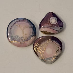 Peut inclure: Trois cabochons lisses et polis présentant des paysages peints à la main. Une pièce circulaire et deux pièces triangulaires affichent des nuances de violet, de rose et de blanc avec des lignes dorées représentant des arbres et une pleine lune.