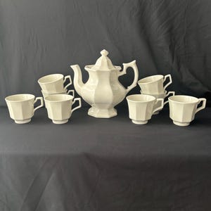 Peut inclure: Un service à thé en céramique blanche composé d'une théière centrale avec couvercle et de six tasses assorties. Chaque pièce présente un design octogonal à facettes distinctif avec une anse. La théière possède un bec verseur courbé.