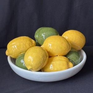 May include: A white ceramic bowl filled with a collection of glossy, realistic-looking ceramic fruit. The arrangement includes several bright yellow lemons and deep green limes, all featuring a smooth, reflective finish and textured surfaces.