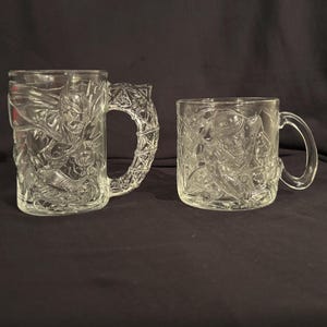 May include: Two clear glass mugs featuring embossed designs. The taller mug displays a caped figure with a textured handle, while the shorter, wider mug features a character in a hat. Both items are made of transparent glass with relief details.